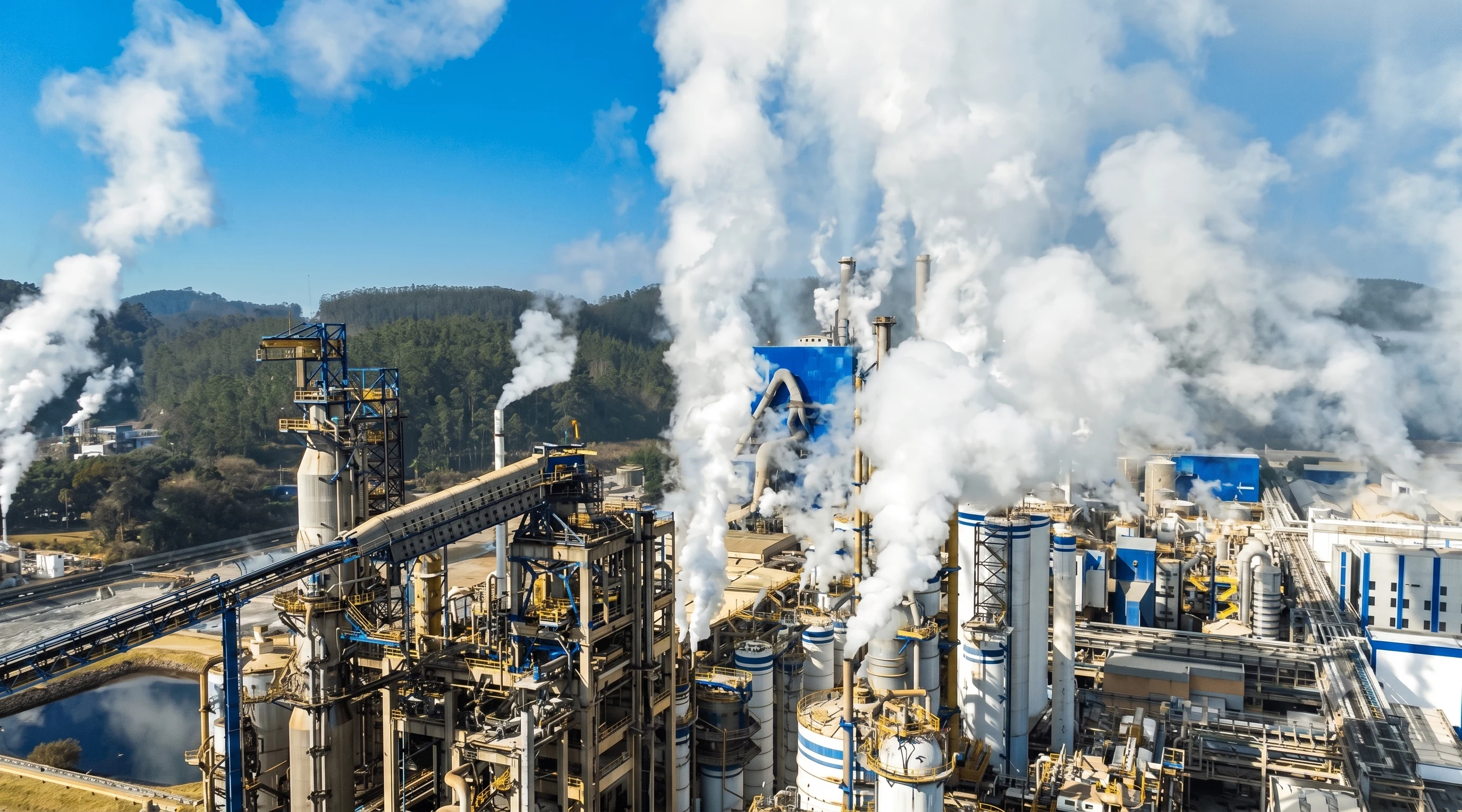 Aerial view of an industrial complex with steam plumes, conveyor belts and forested hills in the background.