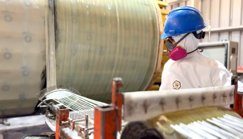 Operario con traje de protección, casco y respirador junto a un cuerpo cilíndrico de FRP en fabricación, con guías de fibra en primer plano.