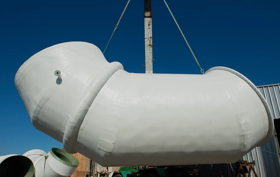 Large-diameter FRP piping elbow suspended from a crane against the sky.