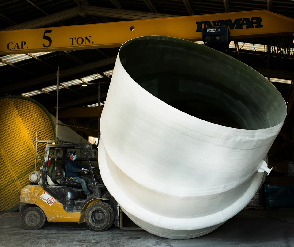 Large-diameter FRP shell course moved by forklift inside a workshop, under an overhead crane.